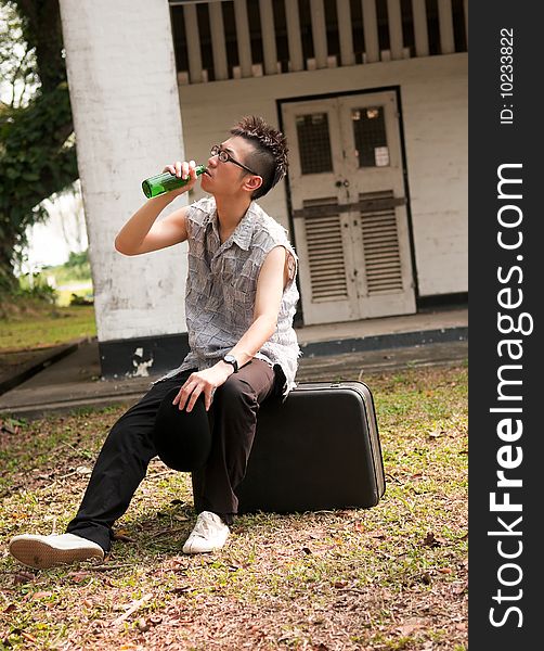 Bespectacled asian man drinking from a bottle and sitting on a suitcase outside the boarded up door of an abandoned apartment. Bespectacled asian man drinking from a bottle and sitting on a suitcase outside the boarded up door of an abandoned apartment