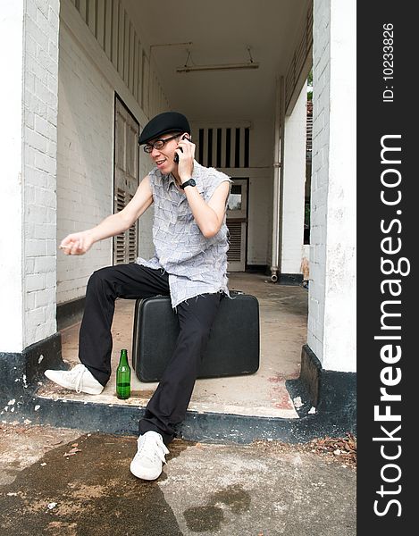 Bespectacled Asian man speaking on the mobile phone while sitting on a suitcase at the end of a corridor of a boarded up and abandoned apartment. Bespectacled Asian man speaking on the mobile phone while sitting on a suitcase at the end of a corridor of a boarded up and abandoned apartment