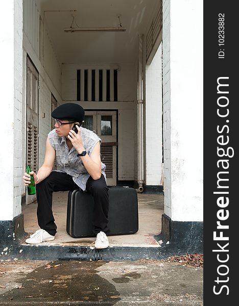 Bespectacled Asian man listening to the mobile phone while sitting on a suitcase at the end of a corridor of a boarded up and abandoned apartment. Bespectacled Asian man listening to the mobile phone while sitting on a suitcase at the end of a corridor of a boarded up and abandoned apartment