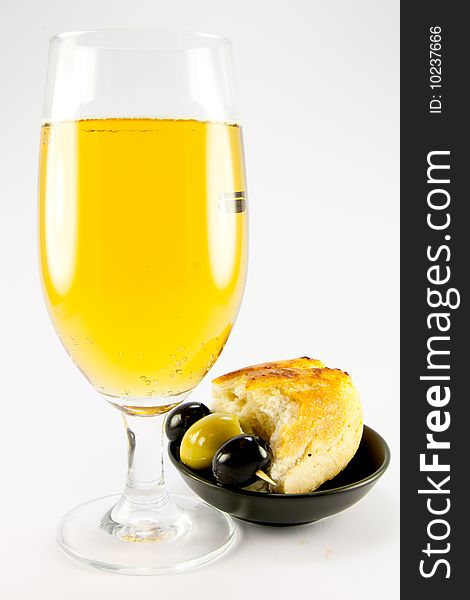 Glass of lager with three green olives and piece of crusty bread in a small black bowl on a plain background. Glass of lager with three green olives and piece of crusty bread in a small black bowl on a plain background