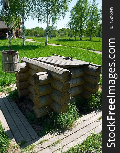 Wooden draw well in the rural area with shadoof and bucket. Green grass around it. Wooden house in background. Wooden draw well in the rural area with shadoof and bucket. Green grass around it. Wooden house in background