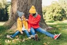 Little Daughter And Her Mother Have Fun Together, Dressed Warm, Sit Near Big Tree On Green Grass, Look At Each Other With Love. Af Royalty Free Stock Photography
