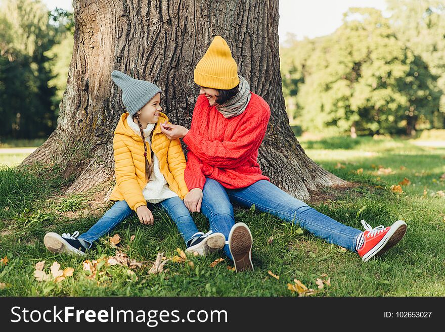 Little daughter and her mother have fun together, dressed warm, sit near big tree on green grass, look at each other with love. Affectionate mom and small female kid have good mood, spend weekends