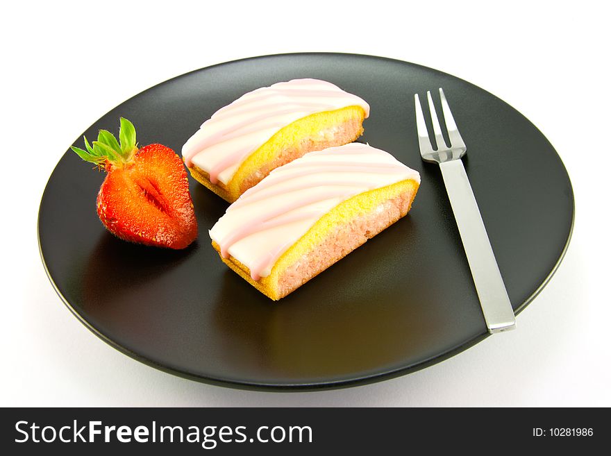 Two pink strawberry sliced cakes with icing on the top with a half strawberry and small fork on a black plate with a white background. Two pink strawberry sliced cakes with icing on the top with a half strawberry and small fork on a black plate with a white background