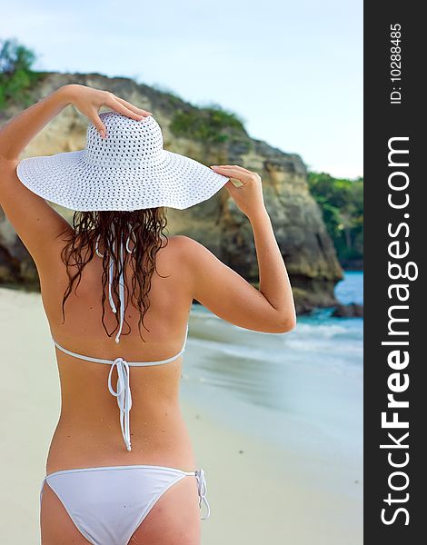 Beautiful brunette with long hair in white hat stands on the sand ocean beach on Bali in Indonesia. Beautiful brunette with long hair in white hat stands on the sand ocean beach on Bali in Indonesia