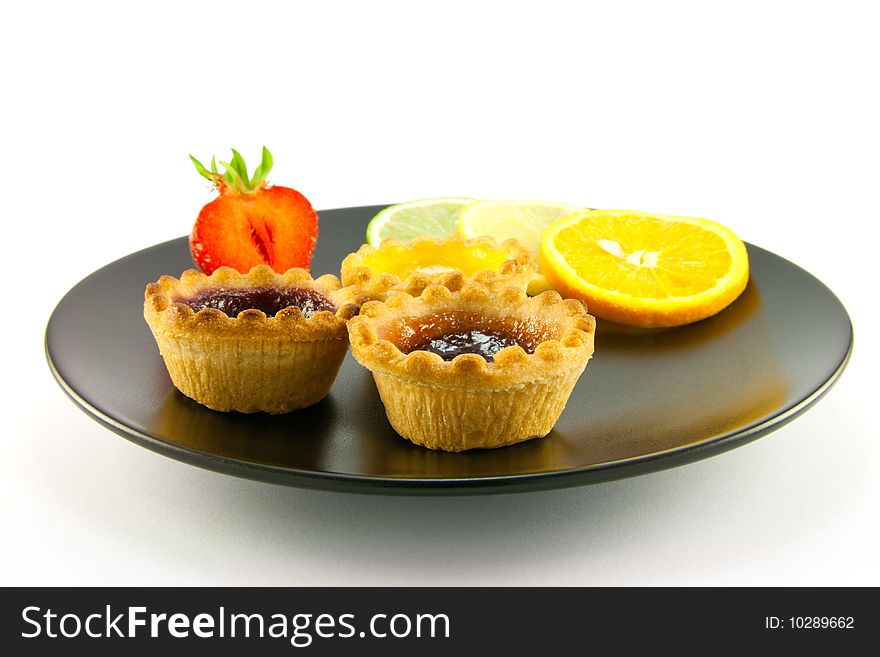Red and yellow small jam tarts with slices of lemon, lime, orange and strawberry on a black plate on a white background. Red and yellow small jam tarts with slices of lemon, lime, orange and strawberry on a black plate on a white background