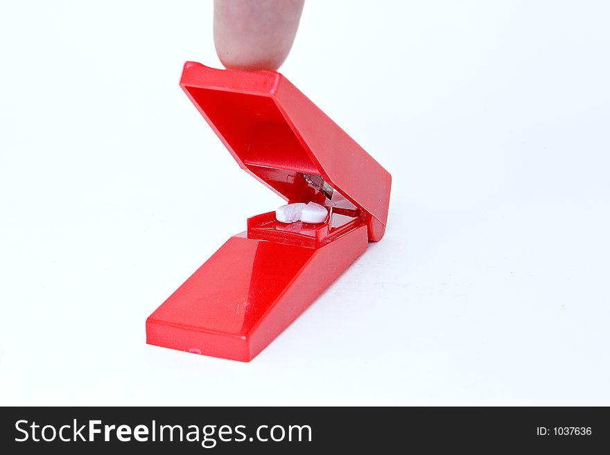A red pill splitter which just split a white pill. The finger which pushed it is shown. White background.