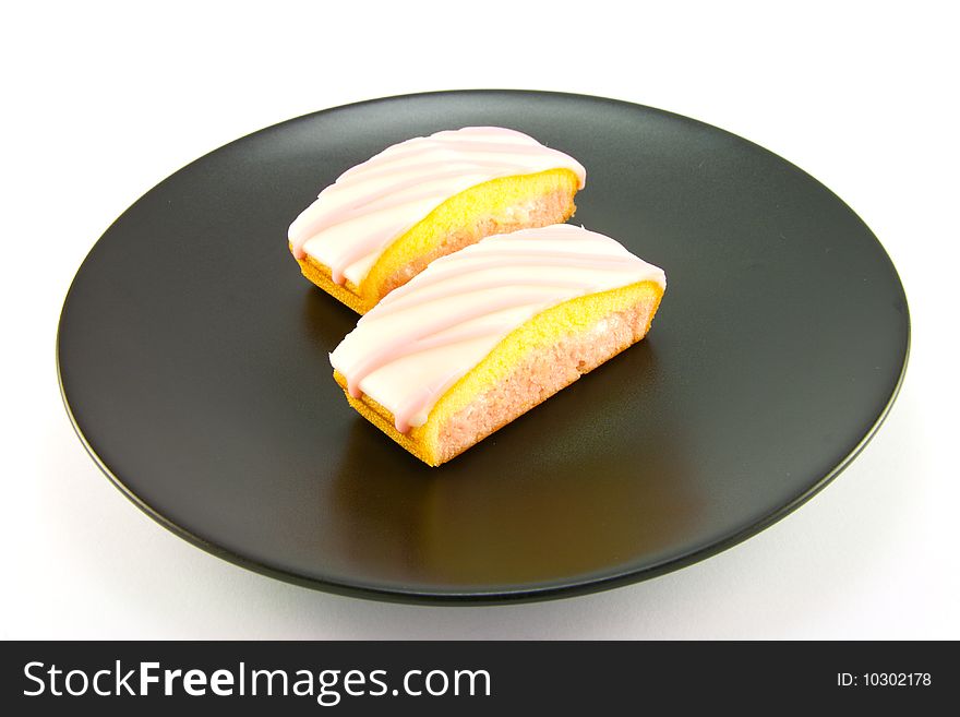 Two strawberry sliced cakes with icing on the top and pink and yellow sponge on a black plate with a white background. Two strawberry sliced cakes with icing on the top and pink and yellow sponge on a black plate with a white background