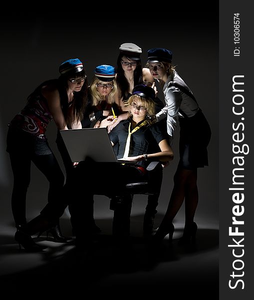 Team of five young women in generic university caps leaning over laptop. Black background, dramatic lighting. Team of five young women in generic university caps leaning over laptop. Black background, dramatic lighting.
