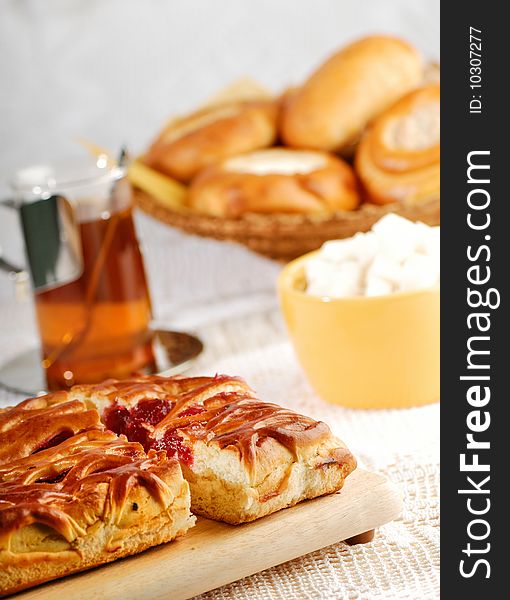 A pie with fruit jam, several patties, sugar in yellow bowl and a cup of tea on the table with white tablecloth. A pie with fruit jam, several patties, sugar in yellow bowl and a cup of tea on the table with white tablecloth