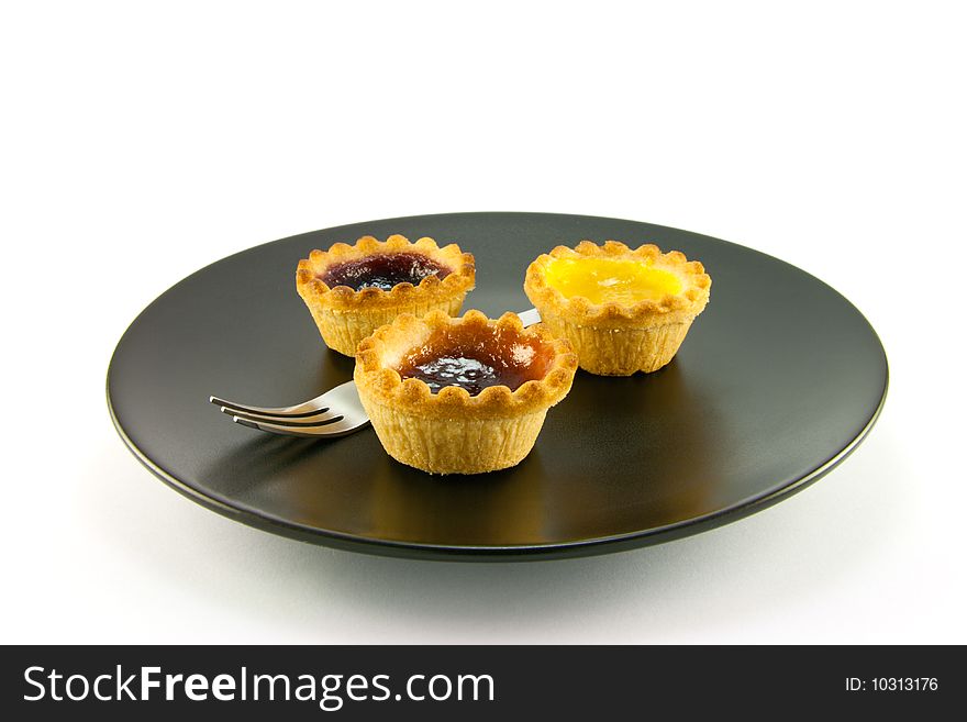 Three red and yellow small pastry jam tarts on a black plate with a fork on a white background. Three red and yellow small pastry jam tarts on a black plate with a fork on a white background