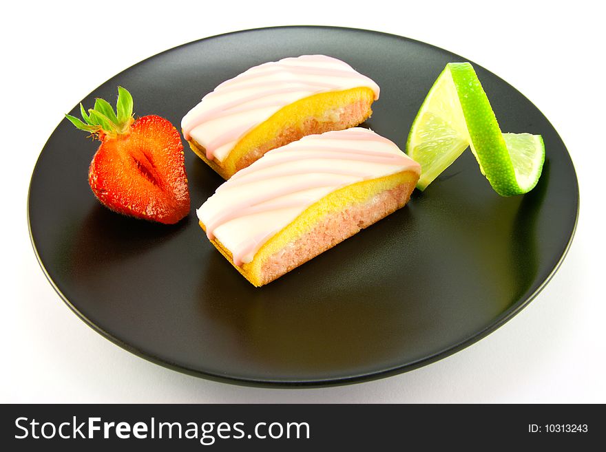 Two pink strawberry sliced cakes with icing on the top with a half strawberry and twist of lime on a black plate on a white background. Two pink strawberry sliced cakes with icing on the top with a half strawberry and twist of lime on a black plate on a white background