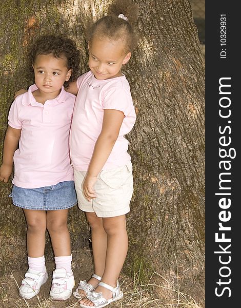 Two Young Ethnic Sisters with Questioning and Mischievous looks on their faces. They have their arms around each other as hold each other while standing against the trunk of a large tree.