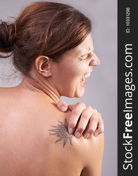 Young woman with sever back pain. She is holding her schoulder. Over grey background. She has a tattoo on her shoulder. Young woman with sever back pain. She is holding her schoulder. Over grey background. She has a tattoo on her shoulder.