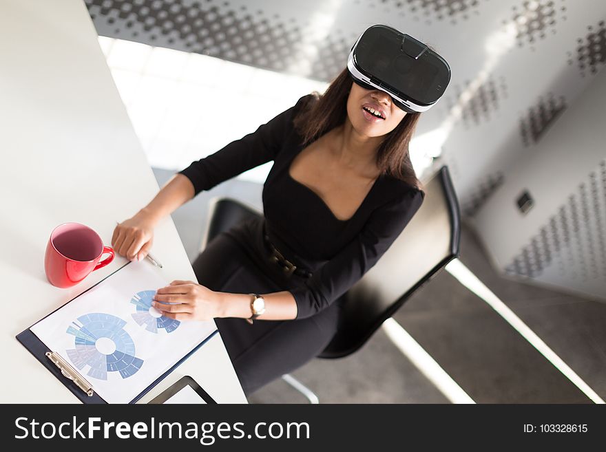 Beauty young brunette afroamerican business lady in black business suit, glasses and costly watch sit at the table in the office, red cup on the table, sit in virtual reality glasses, turned picture. Beauty young brunette afroamerican business lady in black business suit, glasses and costly watch sit at the table in the office, red cup on the table, sit in virtual reality glasses, turned picture