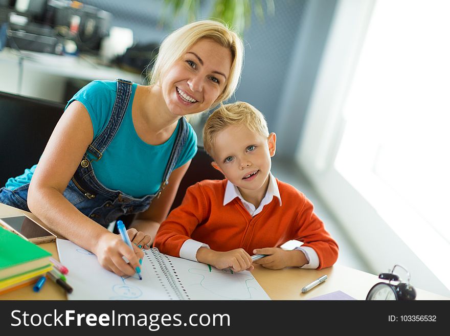 Young blonde caucasian kid in orange shirt and his mother sit at the table with colorful folders and alarm clock and draw together, look at cameranBlurred office on background. Young blonde caucasian kid in orange shirt and his mother sit at the table with colorful folders and alarm clock and draw together, look at cameranBlurred office on background