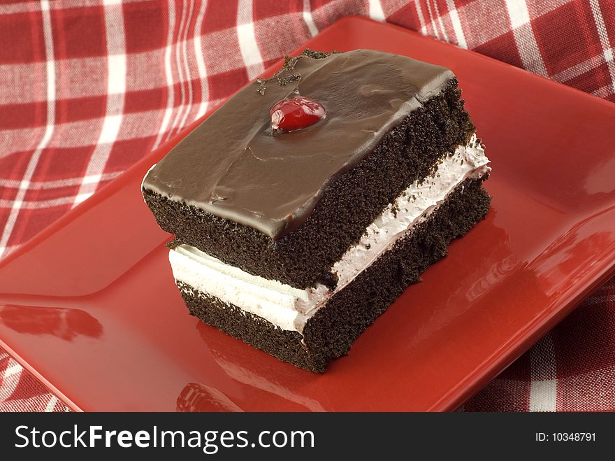 A piece of delicious chocolate layer cake with white frosting in the center layer, and chocolate frosting on top with a cherry, red plate with red plaid tablecloth