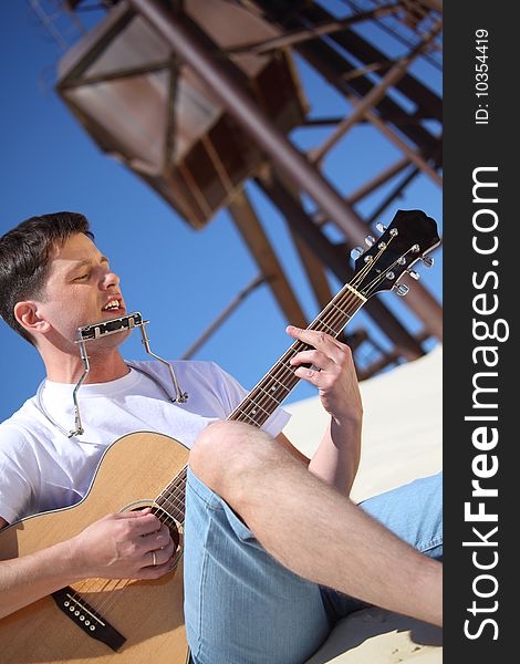 Guy plays guitar and lip accordion sitting on sand. Guy plays guitar and lip accordion sitting on sand