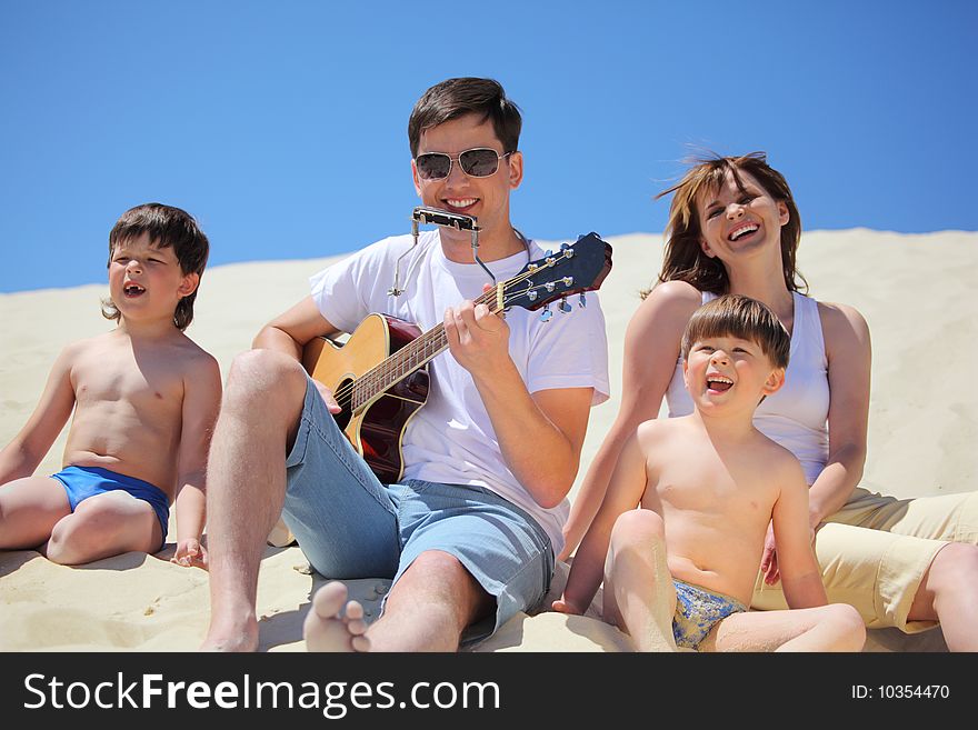 Guy in sunglasses plays guitar and  lip accordion  with children and girl singing song sitting on sand. Guy in sunglasses plays guitar and  lip accordion  with children and girl singing song sitting on sand