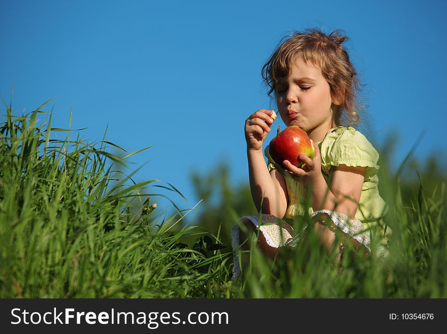 Girl eats red apple in grass against blue sky, closed eyes