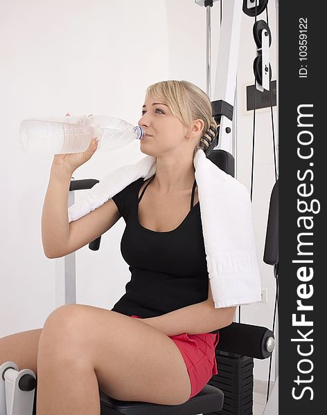 Young blond caucasian woman working out in gym. Smiling model holds bottle with clear drinking water. 3/4 view. Happy facial expression. Model looks aside. Young blond caucasian woman working out in gym. Smiling model holds bottle with clear drinking water. 3/4 view. Happy facial expression. Model looks aside.