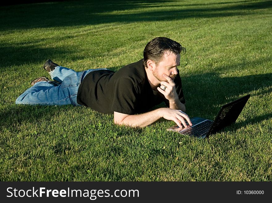 A young Caucasian man works with his laptop computer on the lush green grass. A young Caucasian man works with his laptop computer on the lush green grass.
