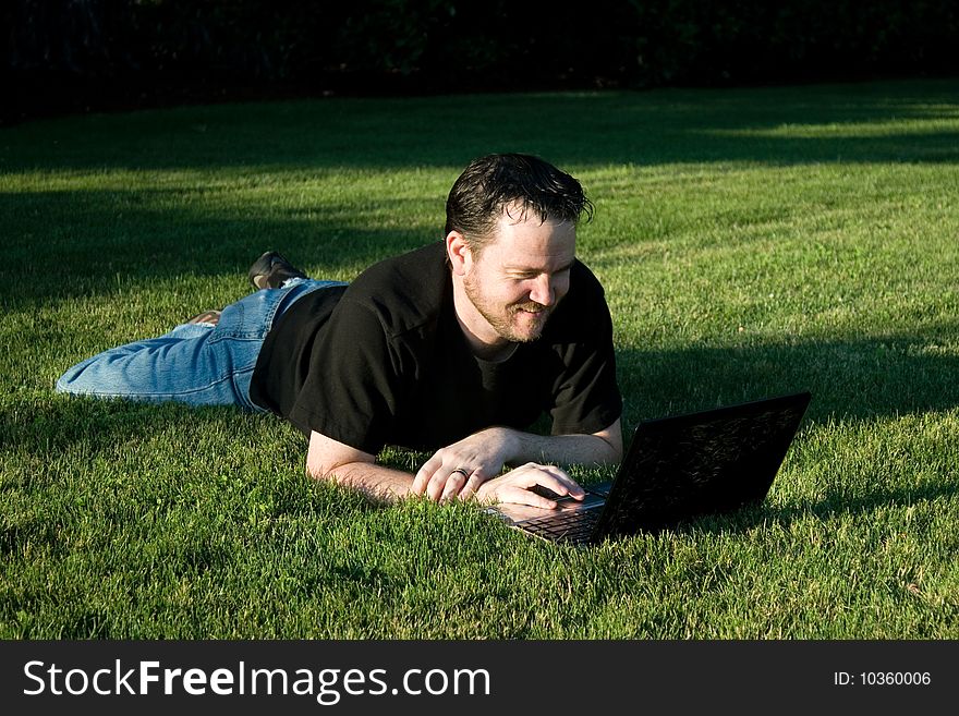 A young Caucasian man works with his laptop computer on the lush green grass. A young Caucasian man works with his laptop computer on the lush green grass.