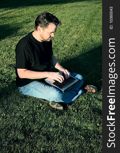 A young Caucasian man works with his laptop computer on the lush green grass. A young Caucasian man works with his laptop computer on the lush green grass.