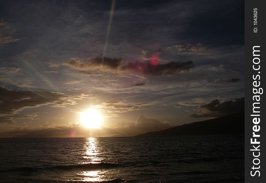 Ocean sunset or sunrise with lens flair and rays, partly cloudy. Hawaii islands off to the side