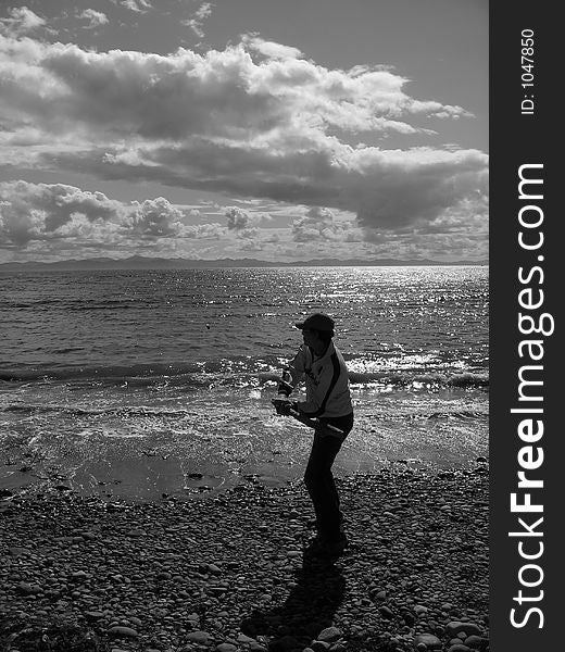 Man. standing at the beach hitting rocks with driftwood. Photo is in black and white with man in center ofpicture and ocean and interesting sky background. Man. standing at the beach hitting rocks with driftwood. Photo is in black and white with man in center ofpicture and ocean and interesting sky background