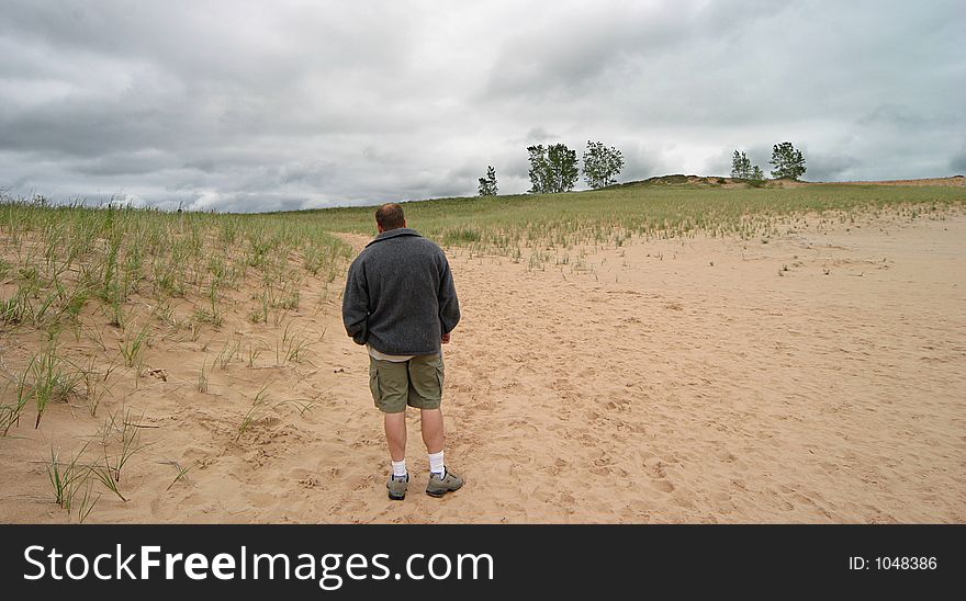 Man standing in a sandy dunes region with some trees and threatening skies ahead. Man standing in a sandy dunes region with some trees and threatening skies ahead.