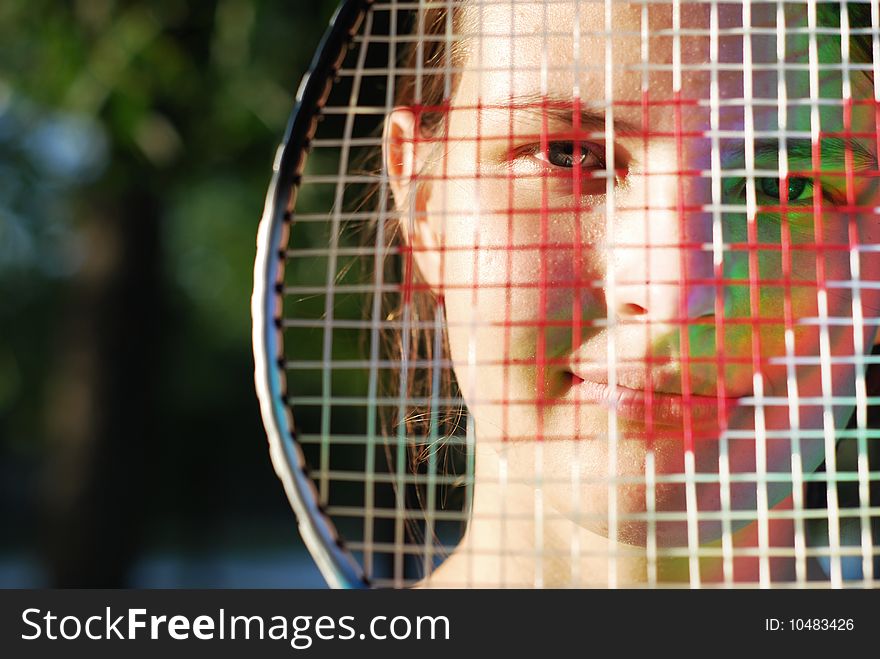 Young beautiful face through the badminton racket; lateral warm natural lighting; there is a colour sunbeam on the cheek in the shadow. Young beautiful face through the badminton racket; lateral warm natural lighting; there is a colour sunbeam on the cheek in the shadow