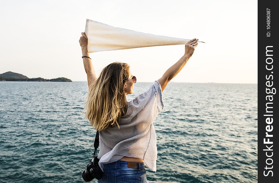 Photo of Woman Wearing White Top, Blue Bottoms and Black Dslr Camera Holding White Textile While Facing the Ocean