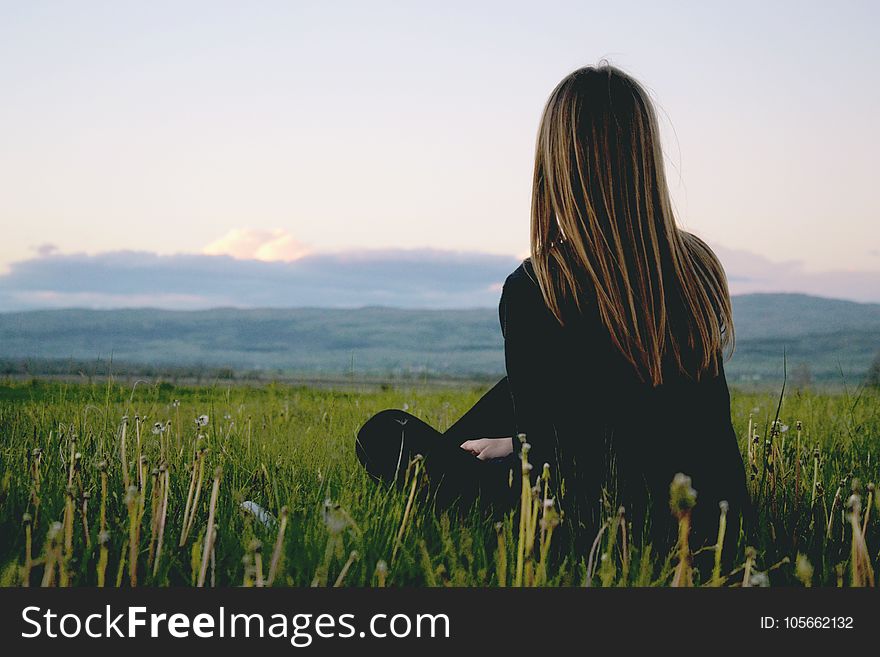 Woman Wearing Black Long Sleeved Shirt Sitting On Green Grass Field Near Mountain Under Cloudy Sky