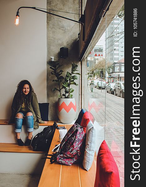 Woman In Gray Zip Up Jacket And Blue Distressed Jeans Sitting On Bench Near Backpack Looking Outside In Room