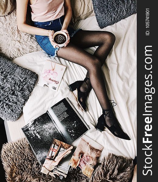 Flatlay Photography Of A Woman Holding White Mug With Black Liquid While Lying On A Bed Surrounded By Fur Pillows And Magazines