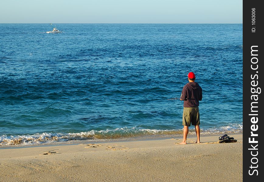 Man with a red cap fishing in the ocean from the beach. The sky is cloudless and blue. The ocean is blue and fairly rough. There is a small amount of surf. A boat is visible not far from the beach. Man with a red cap fishing in the ocean from the beach. The sky is cloudless and blue. The ocean is blue and fairly rough. There is a small amount of surf. A boat is visible not far from the beach.