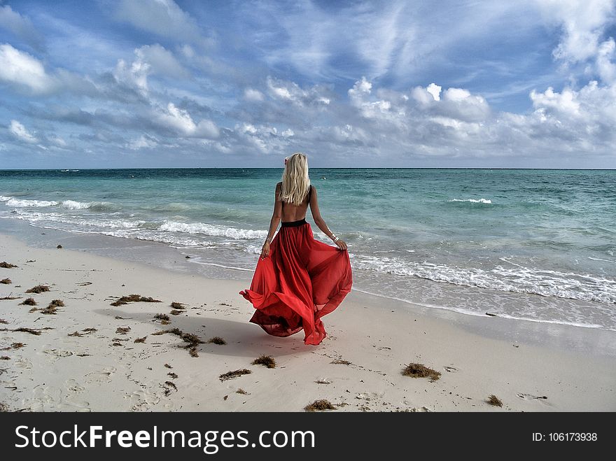Woman Wearing Red Dress Walking on Seashore Under Blue and White Sky