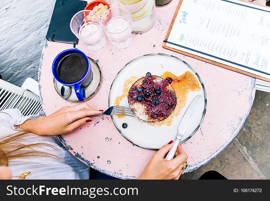 Woman Holding Spoon and Fork With Blackberries on Plate Beside Blue Ceramic Mug on White Wooden Table