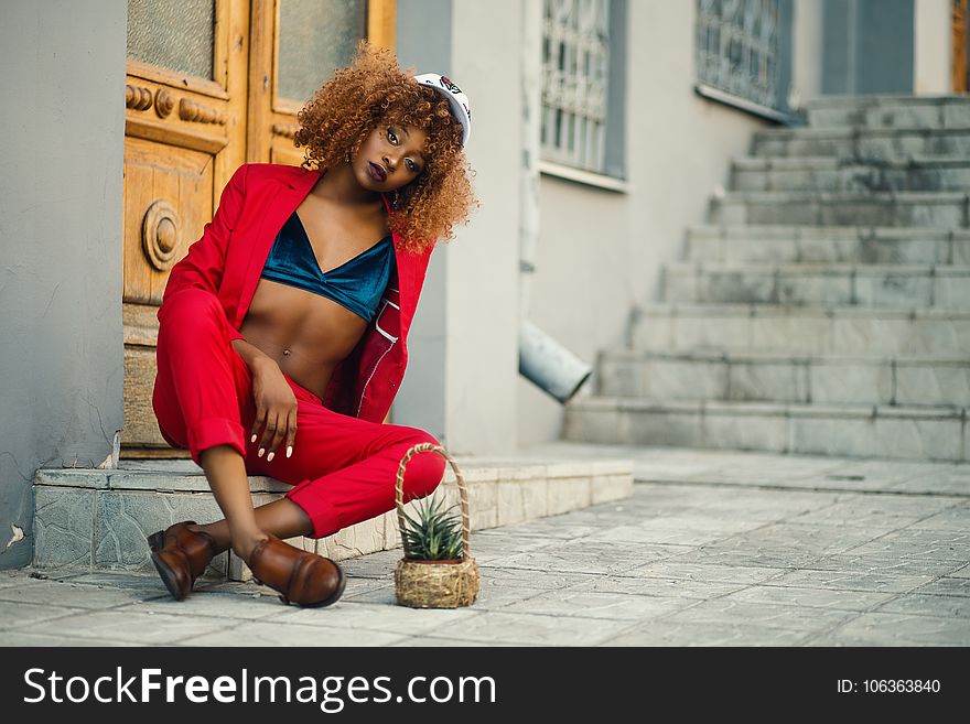 Woman Wearing Red Coat and Pants Sitting Outside Gray Building