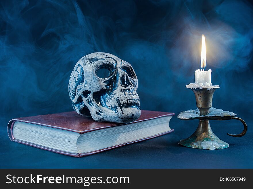 Gothic skull on old book and candle in candlestick on dark and smoked background
