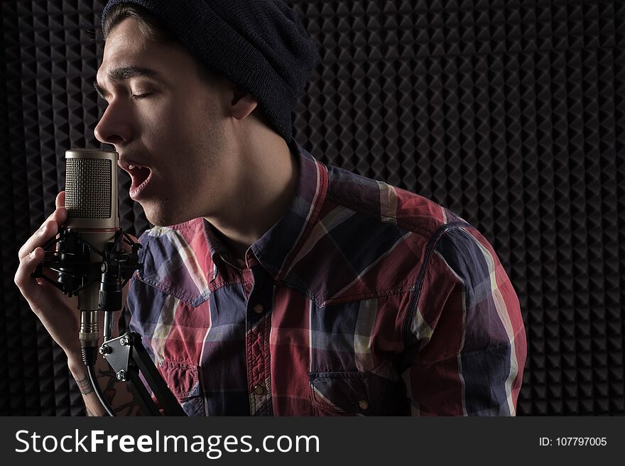 Close-up portrait of emotional young guy singing with his eyes closed in a cap standing in front of the microphone stand holding h
