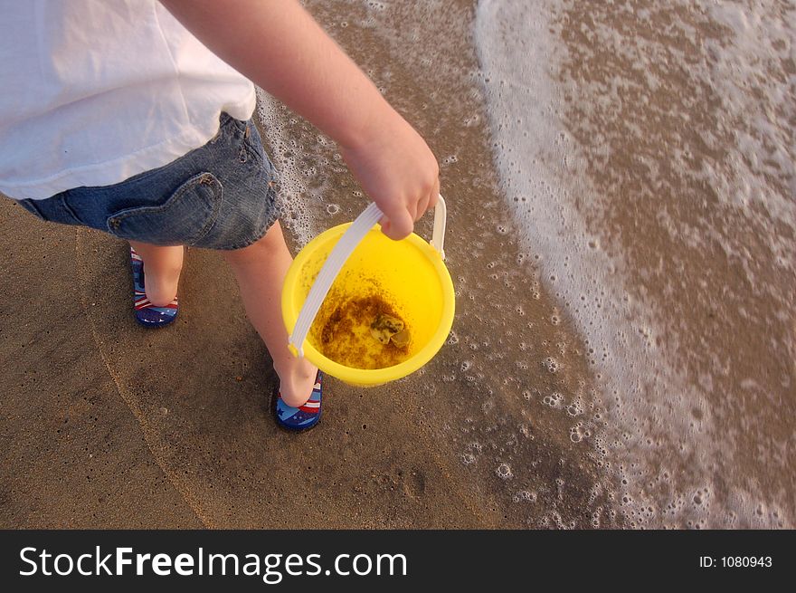 A child just showing her arm and hand carrying a yellow sand bucket on the beach,standing in the foamy waves. A child just showing her arm and hand carrying a yellow sand bucket on the beach,standing in the foamy waves.
