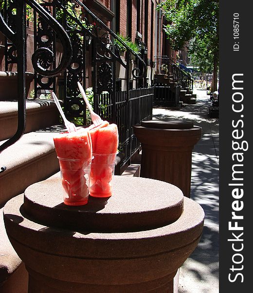 Two tall glasses filled with wattermelon with forks stuck in standing tall on a stoop of a brownstone house. We can see past the stoop all the way up the block. It is a sunny summer morning. Two tall glasses filled with wattermelon with forks stuck in standing tall on a stoop of a brownstone house. We can see past the stoop all the way up the block. It is a sunny summer morning.