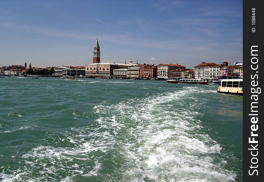 The image shows the view on Venice (Italy) from a ship leaving the city. You see the sea, the waves made from the boat, another ship and the houses of Venice. The tower of San Marco is also on the photo. The sky is blue. The image shows the view on Venice (Italy) from a ship leaving the city. You see the sea, the waves made from the boat, another ship and the houses of Venice. The tower of San Marco is also on the photo. The sky is blue.