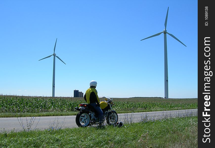 Motorcycle Rider rests while taking in sights of towering windmills. Wearing helmet? Of course. Motorcycle Rider rests while taking in sights of towering windmills. Wearing helmet? Of course.