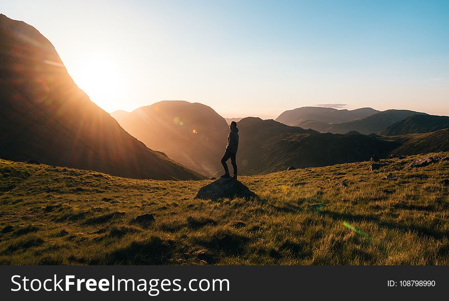 Silhouette Photography of Person Standing on Green Grass in Front of Mountains during Golden Hour