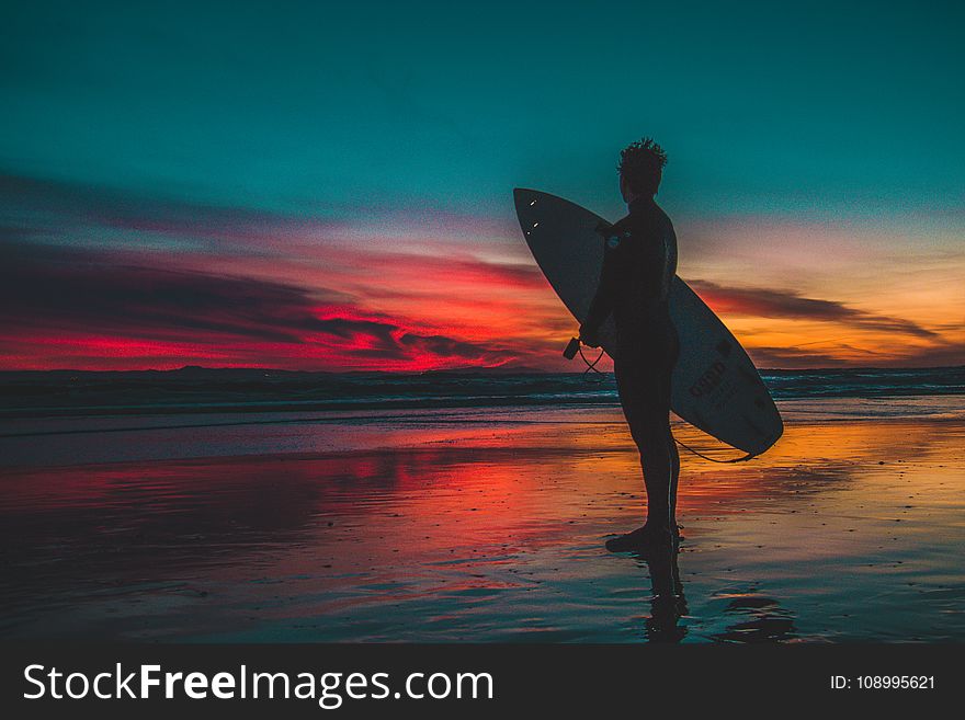 Silhouette of Man in Wet Suit Holding White Surfboard While Standing on Beach during Golden Hour