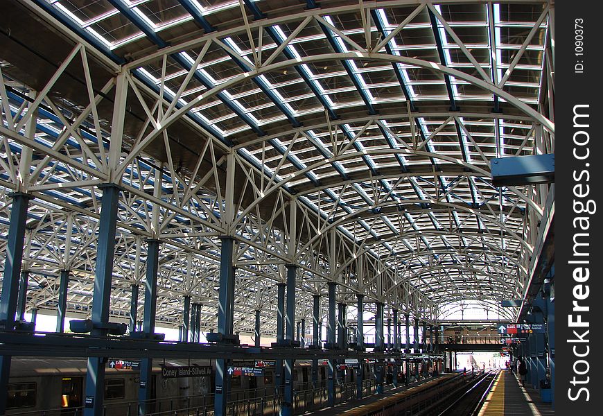 This is the architecture of a large outdoor subway terminal. Very graphic, the cieling and sides are shown in daylight. The ceiling is curved and open with a grid design. The stanchions are painted blue