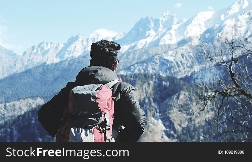 Photography of Man in Black Hooded Jacket and Red Backpack Facing Snow Covered Mountain